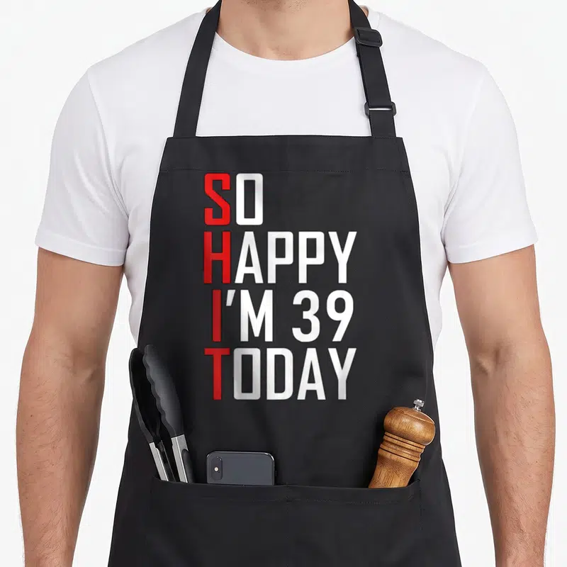 39th joke apron - Stacked red and white text reading SO HAPPY I'M 39 TODAY with vertical SHIT acronym — shown on a black cotton canvas apron against a white background