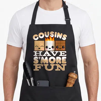 Smore family aprons - Three smiling cartoon s'more ingredients with arched COUSINS text and stacked HAVE S'MORE FUN — shown on a black cotton canvas apron against a white background