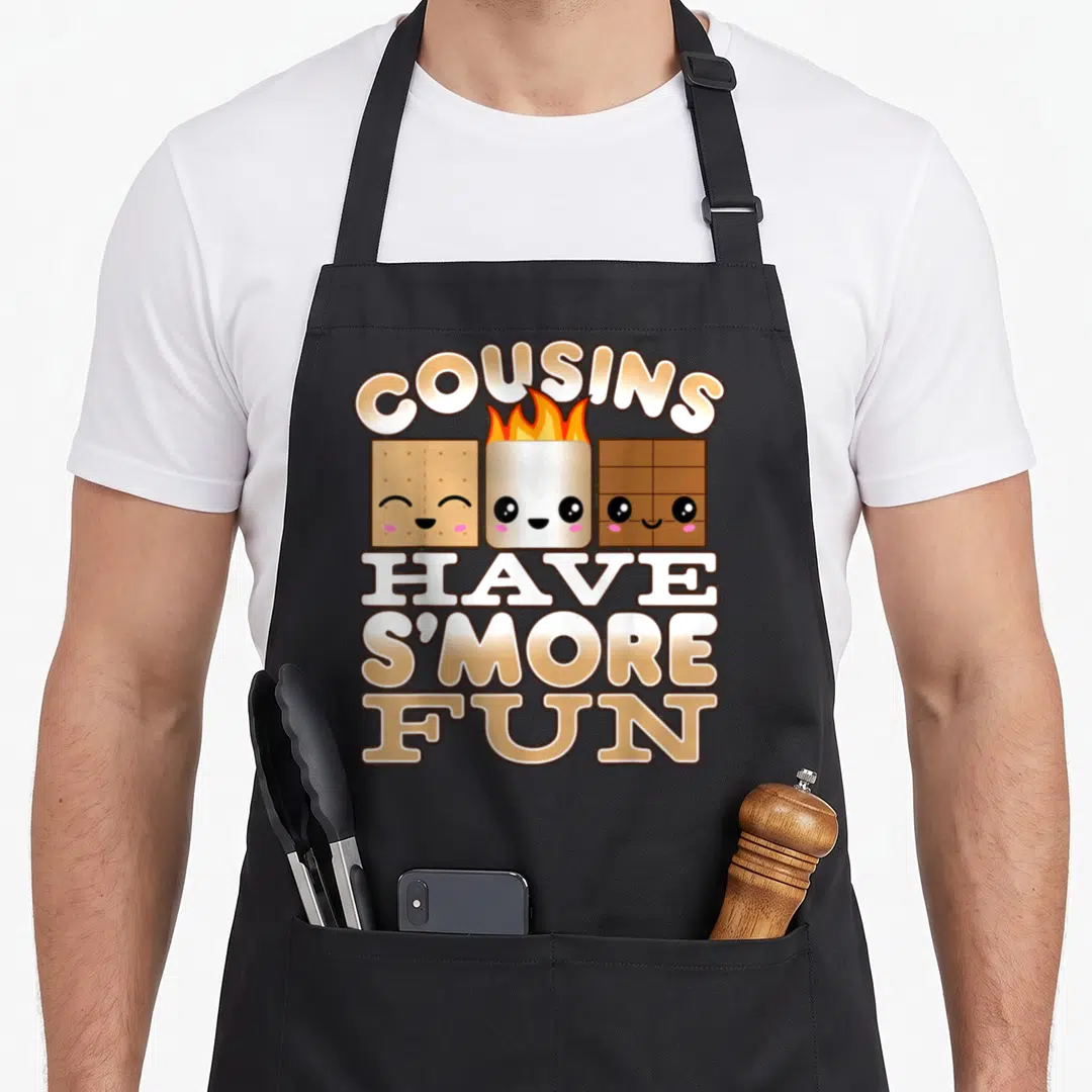 Smore family aprons - Three smiling cartoon s'more ingredients with arched COUSINS text and stacked HAVE S'MORE FUN — shown on a black cotton canvas apron against a white background