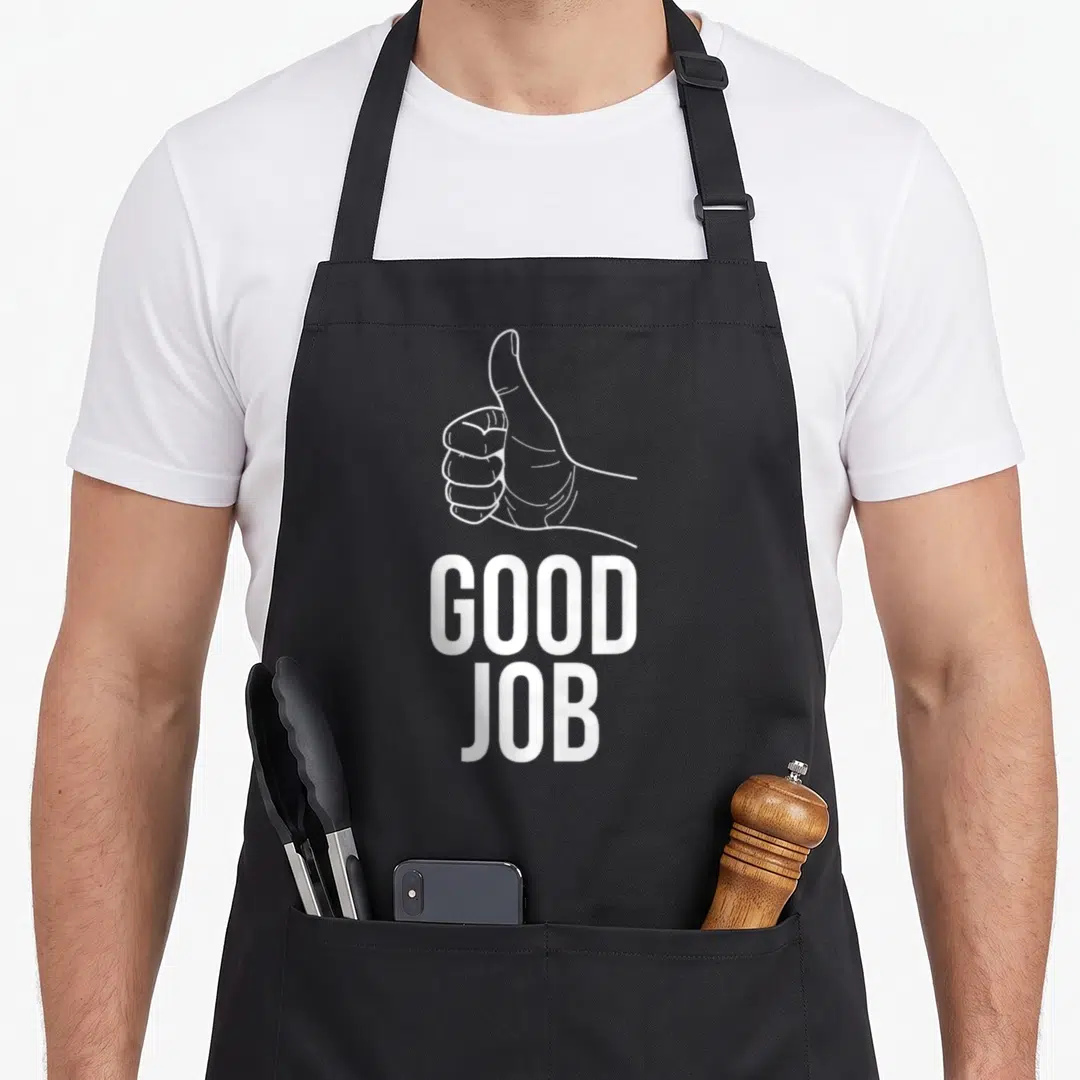Positive thumb apron - White line art hand giving thumbs up above stacked GOOD JOB text on black fabric — shown on a black cotton canvas apron against a white background