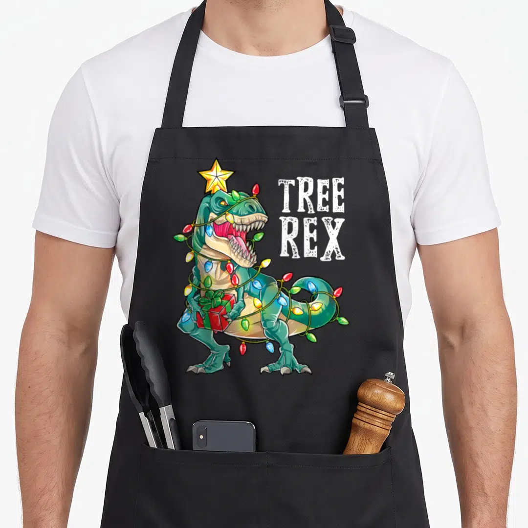 Dinosaur christmas lights apron - Cartoon T-Rex wrapped in multicolor lights holding a red gift with stacked TREE REX — shown on a black cotton canvas apron against a white background