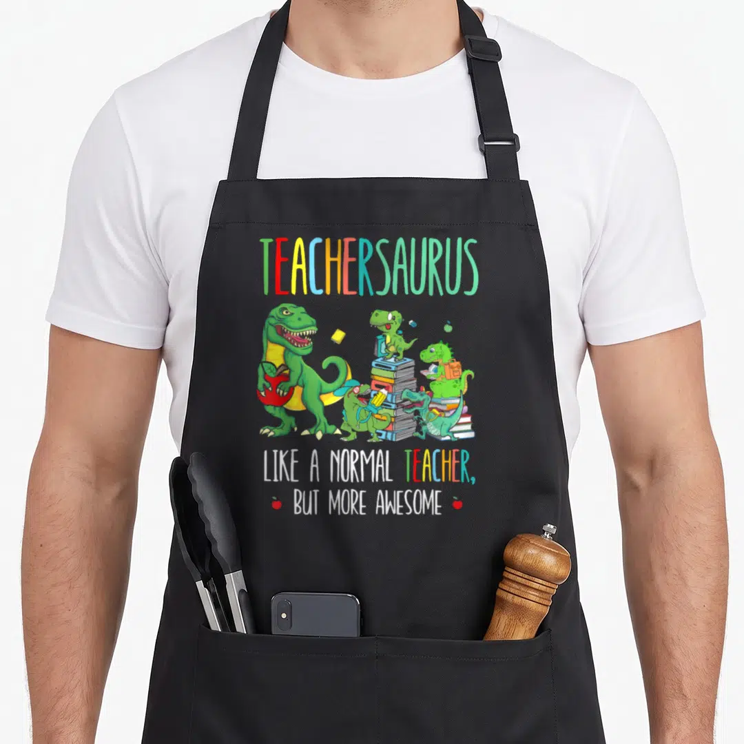 Teachersaurus teacher apron - Colorful cartoon dinosaurs holding books and apples with TEACHERSAURUS text — shown on a black cotton canvas apron against a white background
