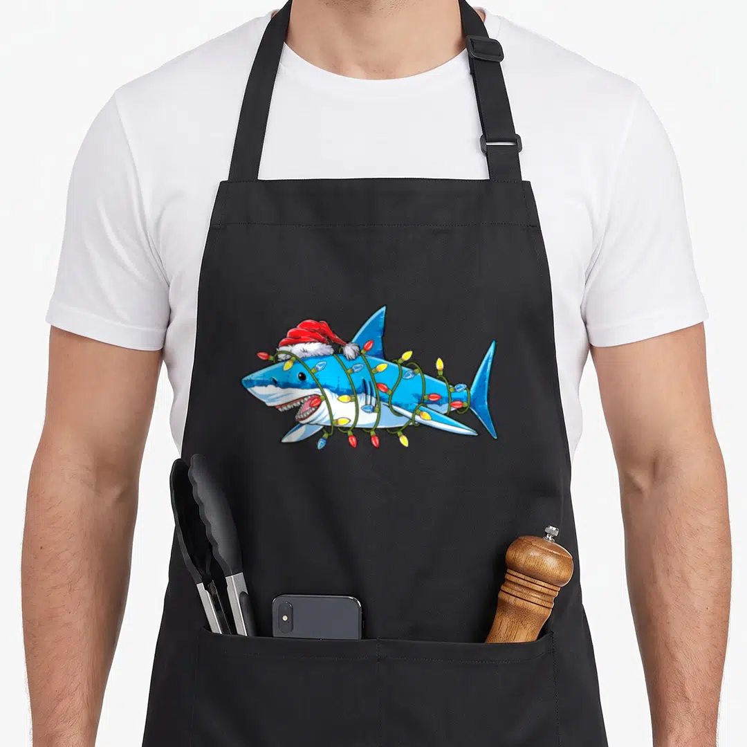 Santa christmas tree apron - Cartoon blue shark wearing red Santa hat wrapped in multicolored Christmas lights — shown on a black cotton canvas apron against a white background
