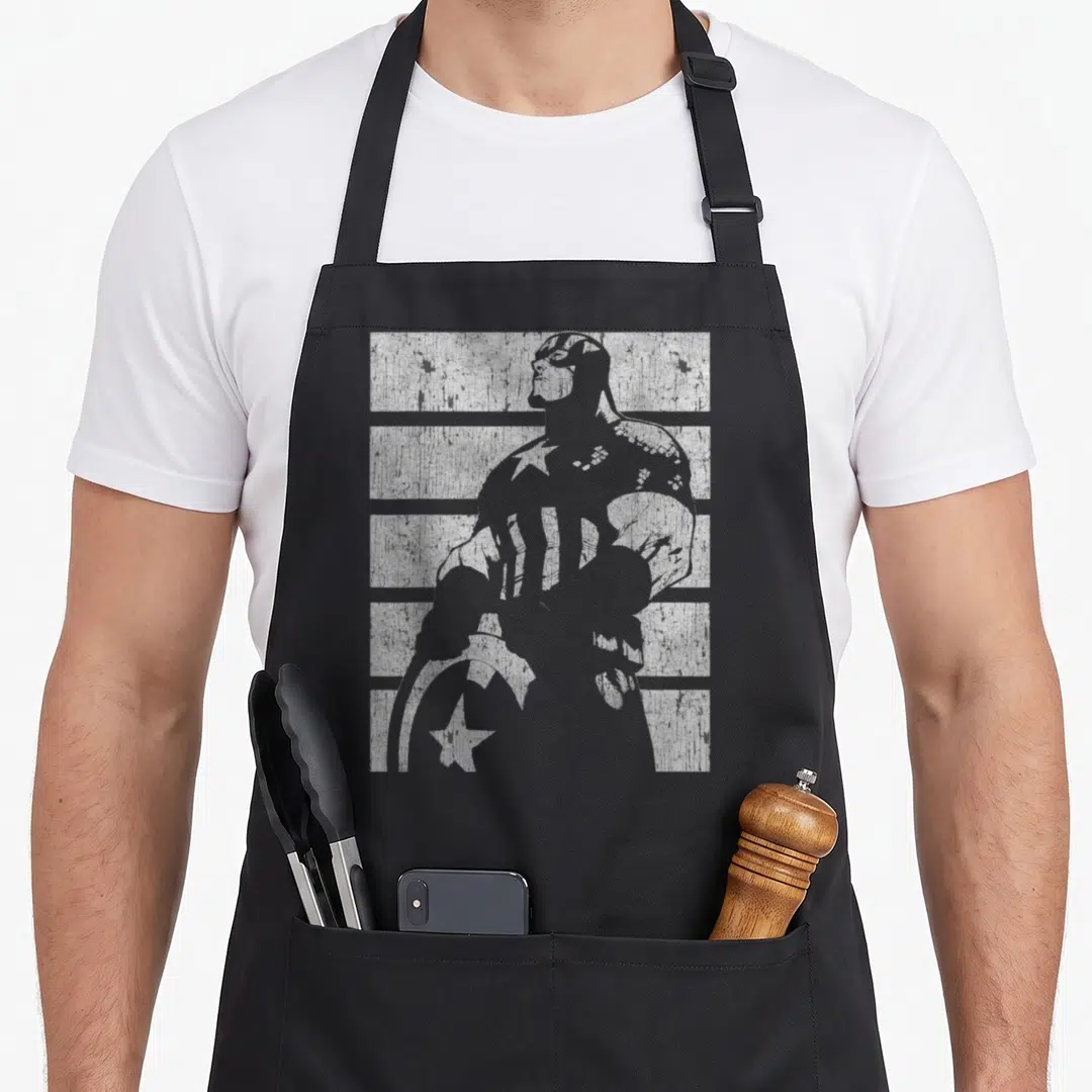Funny silhouette captain apron - Distressed grey silhouette of a masked hero holding a star-spangled shield overlaid — shown on a black cotton canvas apron against a white background