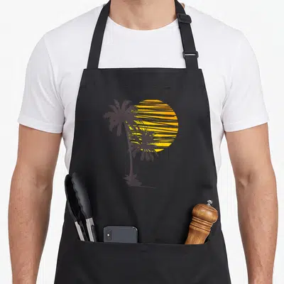Beach holiday apron - Two dark grey palm tree silhouettes against a striped yellow-orange sun — shown on a black cotton canvas apron against a white background