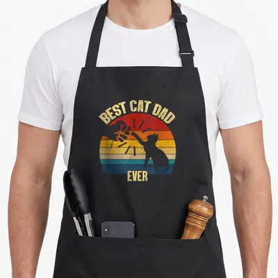 Funny daddy cat dad apron - BEST CAT DAD EVER text over striped sun with cat and thumbs up silhouette — shown on a black cotton canvas apron against a white background