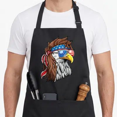 Patriotic bald 4th of july apron - Cartoon bald eagle with brown mullet, star-spangled headband, and flag-patterned — shown on a black cotton canvas apron against a white background