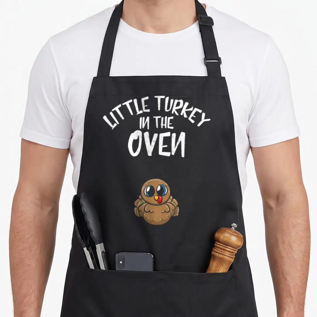 Oven funny turkey apron - LITTLE TURKEY IN THE OVEN text above brown cartoon turkey with blue eyes — shown on a black cotton canvas apron against a white background