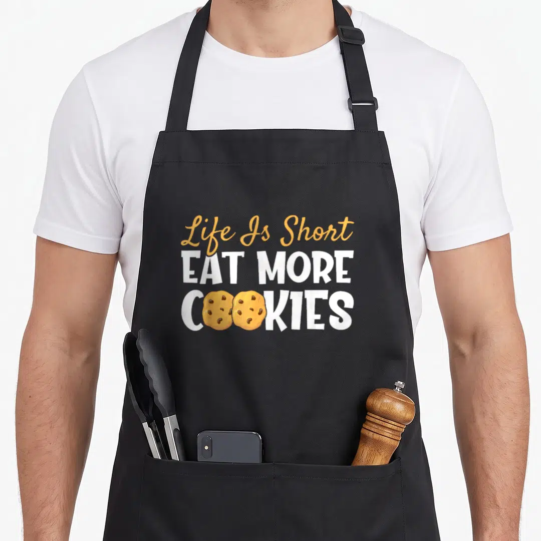 Eat cookies apron - Life Is Short EAT MORE COOKIES text with two tan cookies replacing O letters in white font — shown on a black cotton canvas apron against a white background