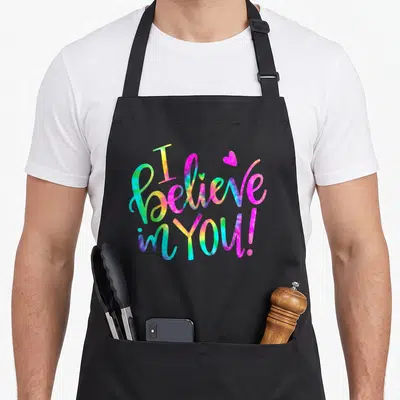 Tie test kitchen apron - Cursive I believe in you text in pink blue yellow and green with a small heart — shown on a black cotton canvas apron against a white background