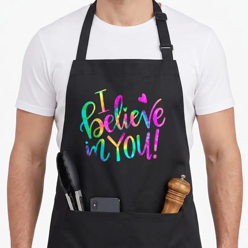 Tie test kitchen apron - Cursive I believe in you text in pink blue yellow and green with a small heart — shown on a black cotton canvas apron against a white background