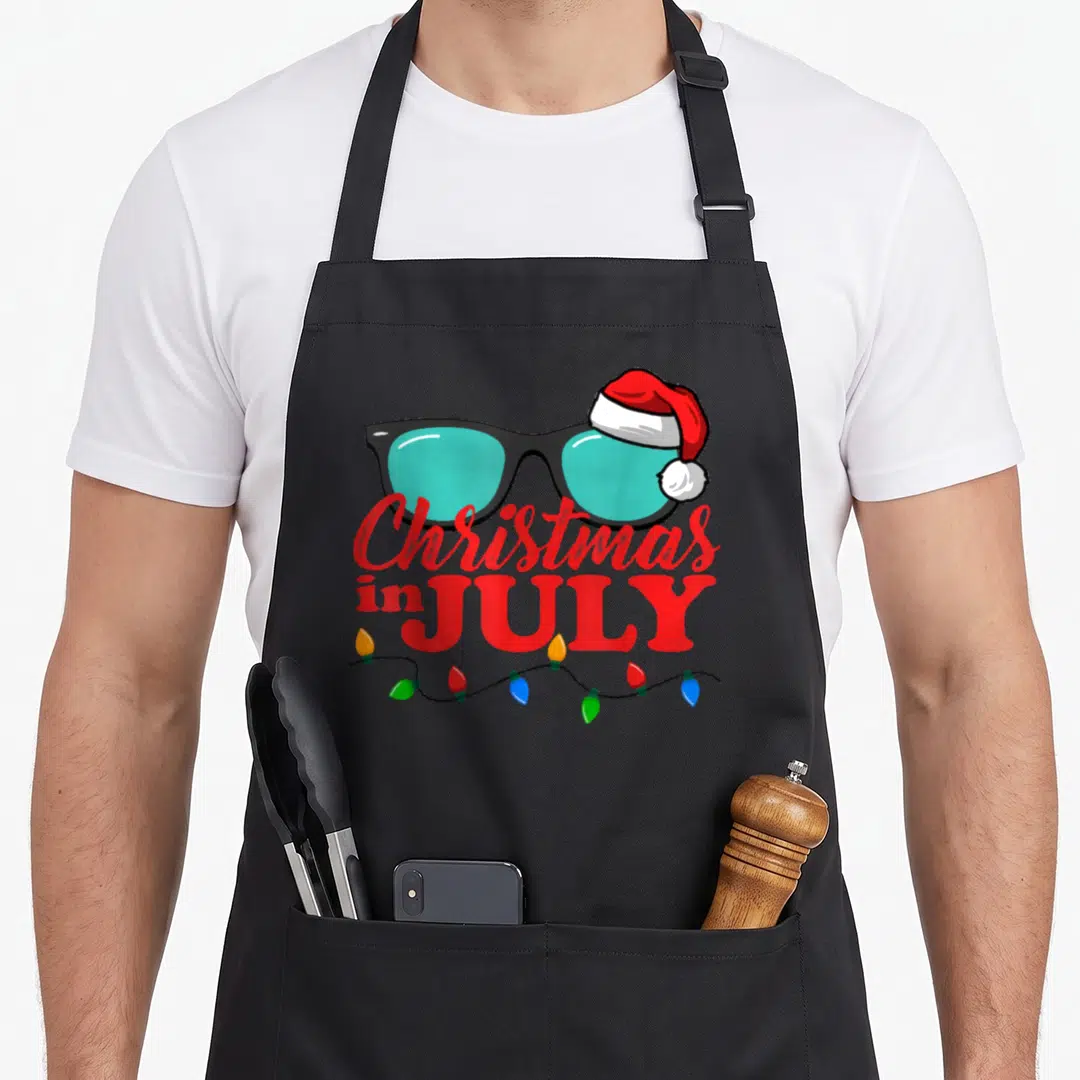 July santa aprons - Sunglasses with red Santa hat, red text, and colorful light bulbs on black wire — shown on a black cotton canvas apron against a white background