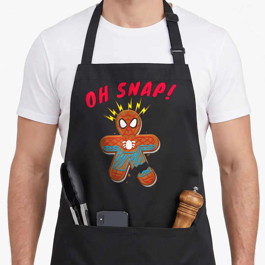 Cookie snap gingerbread apron - Cartoon gingerbread man with spider mask, broken leg, red OH SNAP text, and lightning — shown on a black cotton canvas apron against a white background