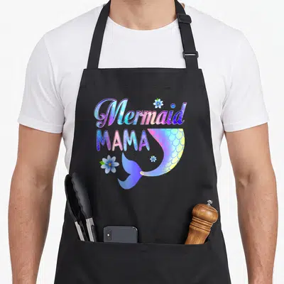 Funny mermaid mama apron - Mermaid MAMA text with gradient tail and three blue flowers in cartoon style — shown on a black cotton canvas apron against a white background