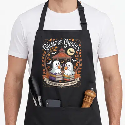 Ghoul gilmore girls apron - Two cartoon ghosts on book stacks holding mugs under GILMORE GHOULS text and gazebo — shown on a black cotton canvas apron against a white background
