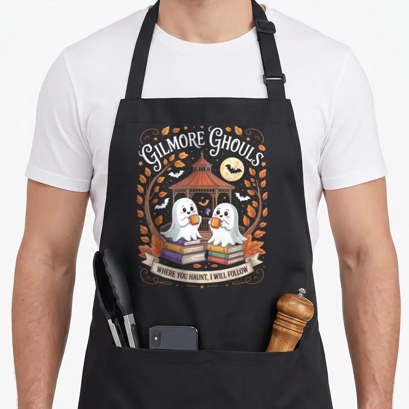 Ghoul gilmore girls apron - Two cartoon ghosts on book stacks holding mugs under GILMORE GHOULS text and gazebo — shown on a black cotton canvas apron against a white background