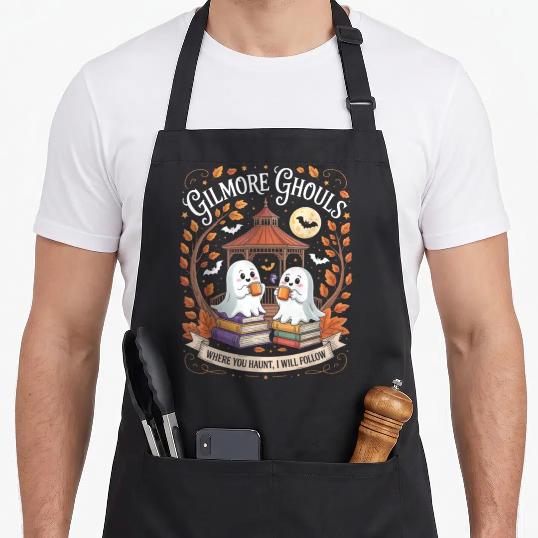 Ghoul gilmore girls apron - Two cartoon ghosts on book stacks holding mugs under GILMORE GHOULS text and gazebo — shown on a black cotton canvas apron against a white background