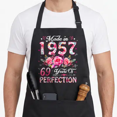 69th vintage floral apron - Stacked Made in 1957 text with pink and white watercolor flowers and small hearts — shown on a black cotton canvas apron against a white background