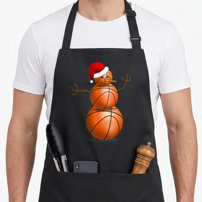 Basketball snowman apron - Three orange basketballs stacked as a snowman with a red Santa hat, stick arms, and carrot — shown on a black cotton canvas apron against a white background