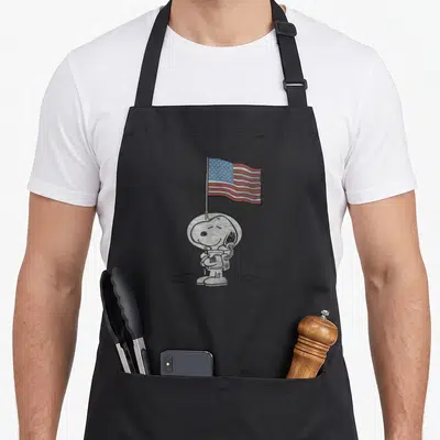 Funny boy astronaut apron - Snoopy in grey astronaut suit holding red white blue flag on grey moon surface line art — shown on a black cotton canvas apron against a white background