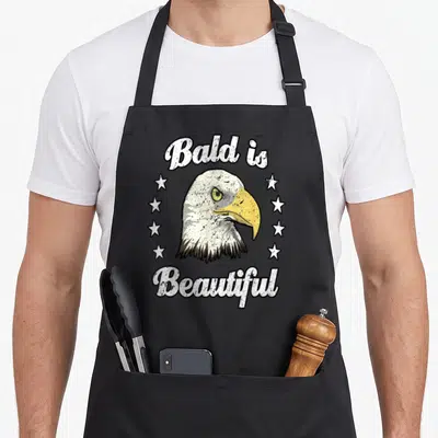 Funny bald beautiful apron - Bald eagle head with arched text and vertical star rows in distressed white, yellow — shown on a black cotton canvas apron against a white background