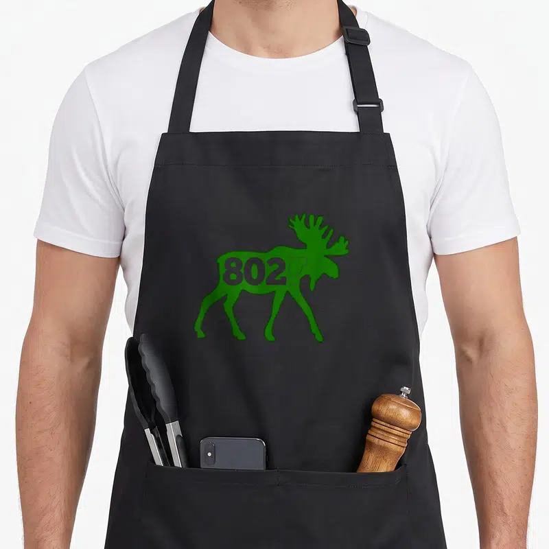 Moose vermont apron - Green moose silhouette with 802 and Vermont state map printed inside — shown on a black cotton canvas apron against a white background
