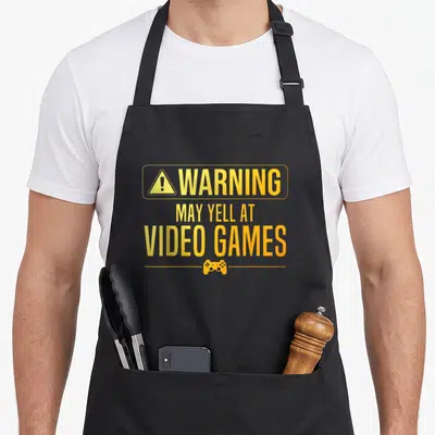 Video nerd apron - Yellow warning sign text and game controller icon on grey background — shown on a black cotton canvas apron against a white background