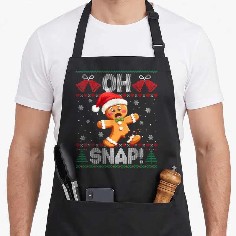 Gingerbread xmas apron - Cartoon gingerbread man with broken leg and OH SNAP text surrounded by knit hearts and trees — shown on a black cotton canvas apron against a white background