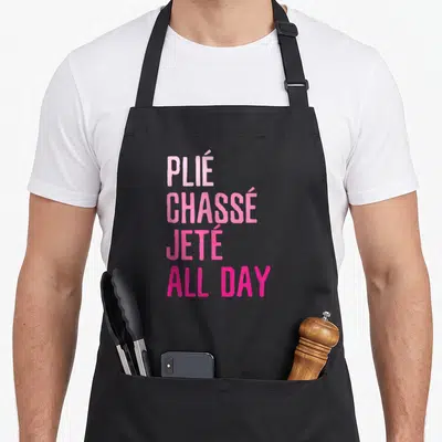 Plie dancing apron - Stacked pink and magenta text reading PLIE CHASSE JETE ALL DAY — shown on a black cotton canvas apron against a white background