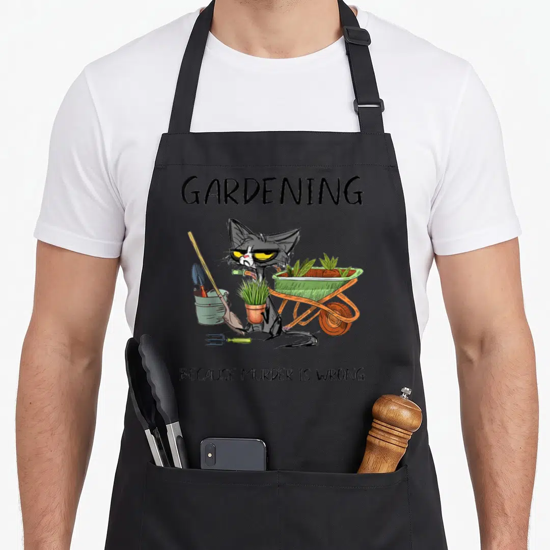 Gardening cat apron - Grumpy cartoon cat holding a plant surrounded by a wheelbarrow and shovel with dark humor text — shown on a black cotton canvas apron against a white background