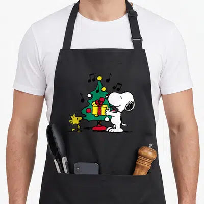 Woodstock christmas tree apron - Snoopy and Woodstock beside a green Christmas tree with musical notes and a yellow — shown on a black cotton canvas apron against a white background