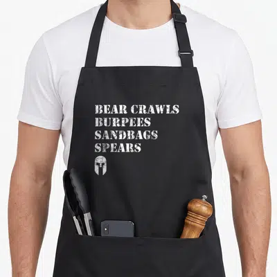 Crawl bear apron - White distressed text reading Bear Crawls Burpees Sandbags Spears above a Spartan helmet icon — shown on a black cotton canvas apron against a white background