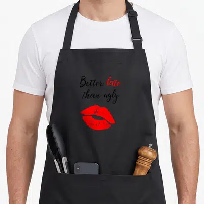 Better ugly apron - Better late than ugly script text above pair of red lips — shown on a black cotton canvas apron against a white background
