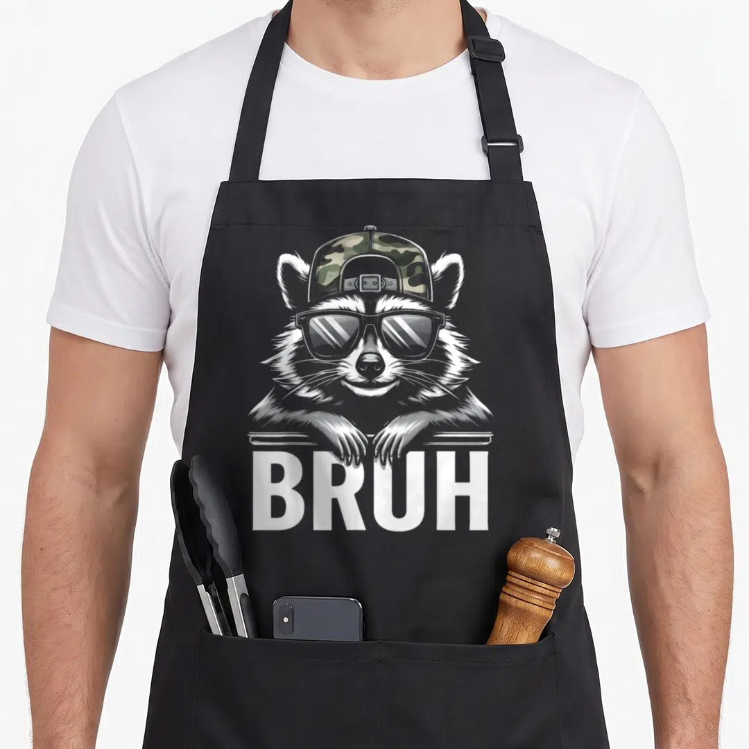 Bruh camo apron - Cartoon raccoon in camo cap and sunglasses leaning over ledge above white BRUH text — shown on a black cotton canvas apron against a white background