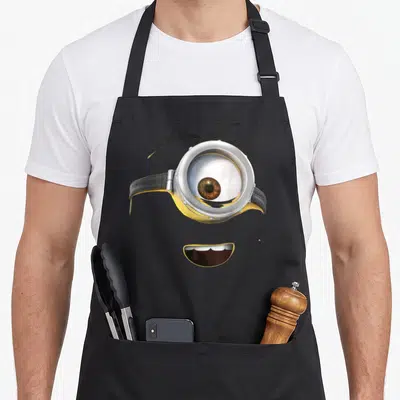 Stuart face apron - Close-up of a yellow minion face with a single silver goggle and smiling mouth — shown on a black cotton canvas apron against a white background