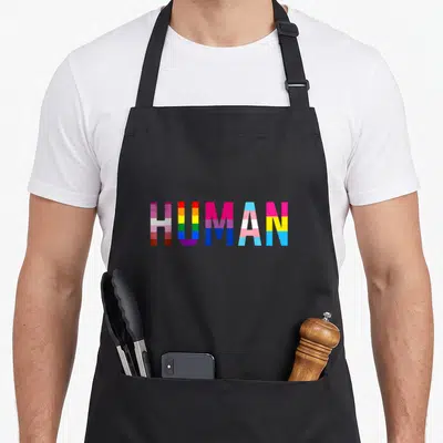 Human rainbow apron - HUMAN text filled with pink, yellow, blue, purple, and red pride flag stripes — shown on a black cotton canvas apron against a white background