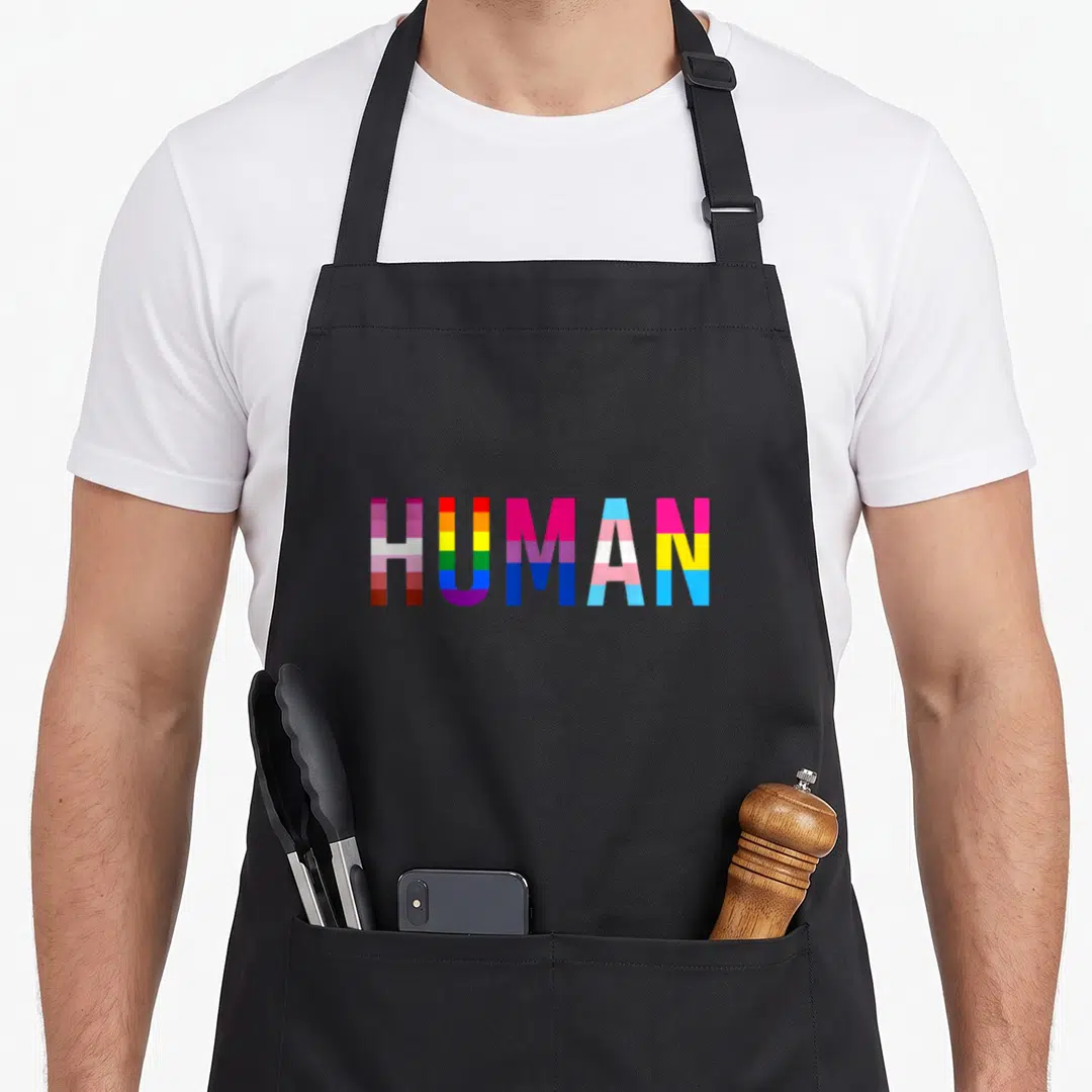 Human rainbow apron - HUMAN text filled with pink, yellow, blue, purple, and red pride flag stripes — shown on a black cotton canvas apron against a white background