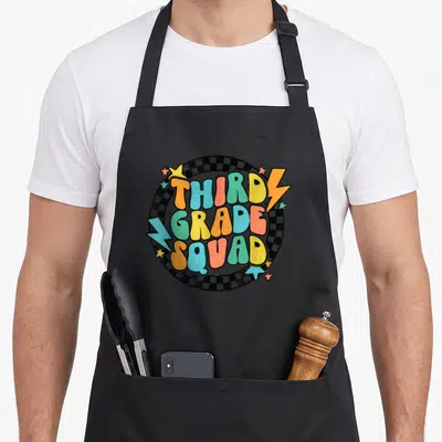 Groovy third teacher apron - Third Grade Squad text over checkerboard circle with colorful lightning bolts and stars — shown on a black cotton canvas apron against a white background