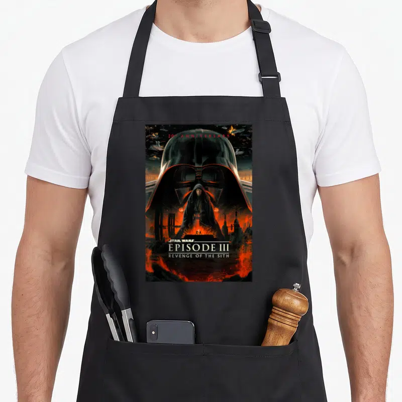 20th anniversary apron - Anakin Skywalker stands before Darth Vader helmet on lava landscape with 20th anniversary text — shown on a black cotton canvas apron against a white background
