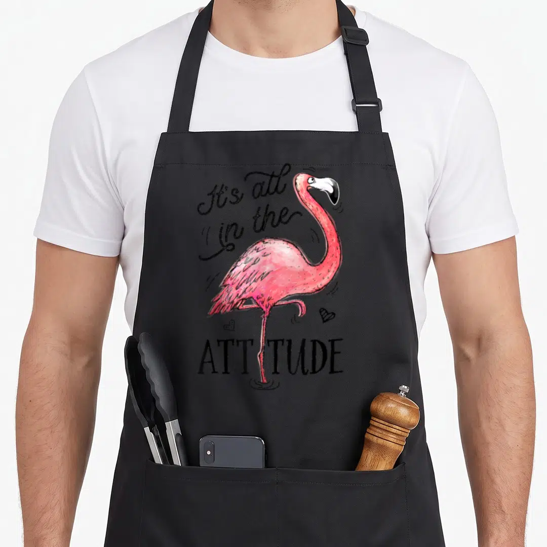Attitude pink flamingo apron - Pink cartoon flamingo standing on one leg with black text It's all in the ATTITUDE — shown on a black cotton canvas apron against a white background