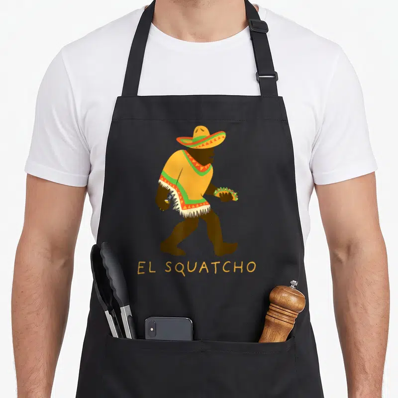Squatcho beer apron - Cartoon bigfoot in sombrero holding taco and bottle with EL SQUATCHO text below — shown on a black cotton canvas apron against a white background
