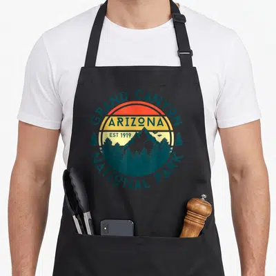 National park canyon apron - Retro circular badge with GRAND CANYON ARIZONA text, striped sun, mountain, and pine trees — shown on a black cotton canvas apron against a white background