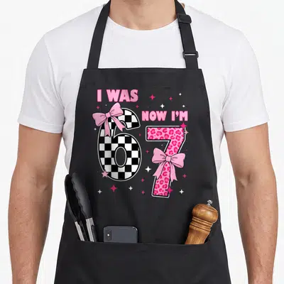 7th bow apron - Checkerboard six and leopard seven with pink bows and stars under I WAS NOW I'M text — shown on a black cotton canvas apron against a white background