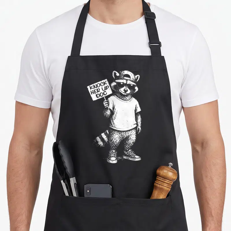 Transfer raccoon dad apron - White line art raccoon wearing sunglasses holding a KNOCK HER UP DOC sign — shown on a black cotton canvas apron against a white background