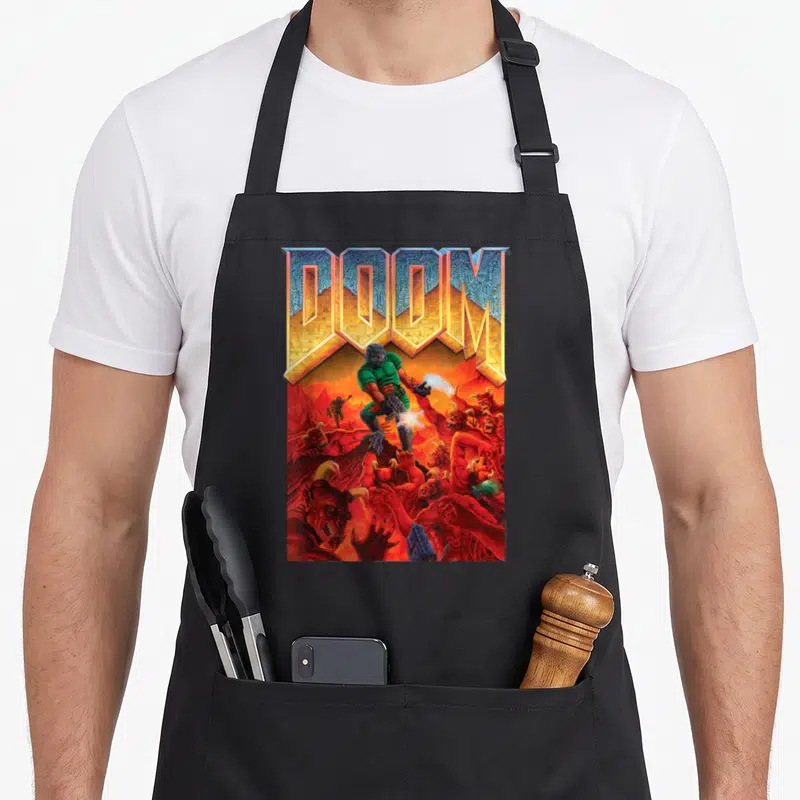 Doom full cover apron - Doomguy fighting red demons below large metallic DOOM text in a fiery landscape — shown on a black cotton canvas apron against a white background