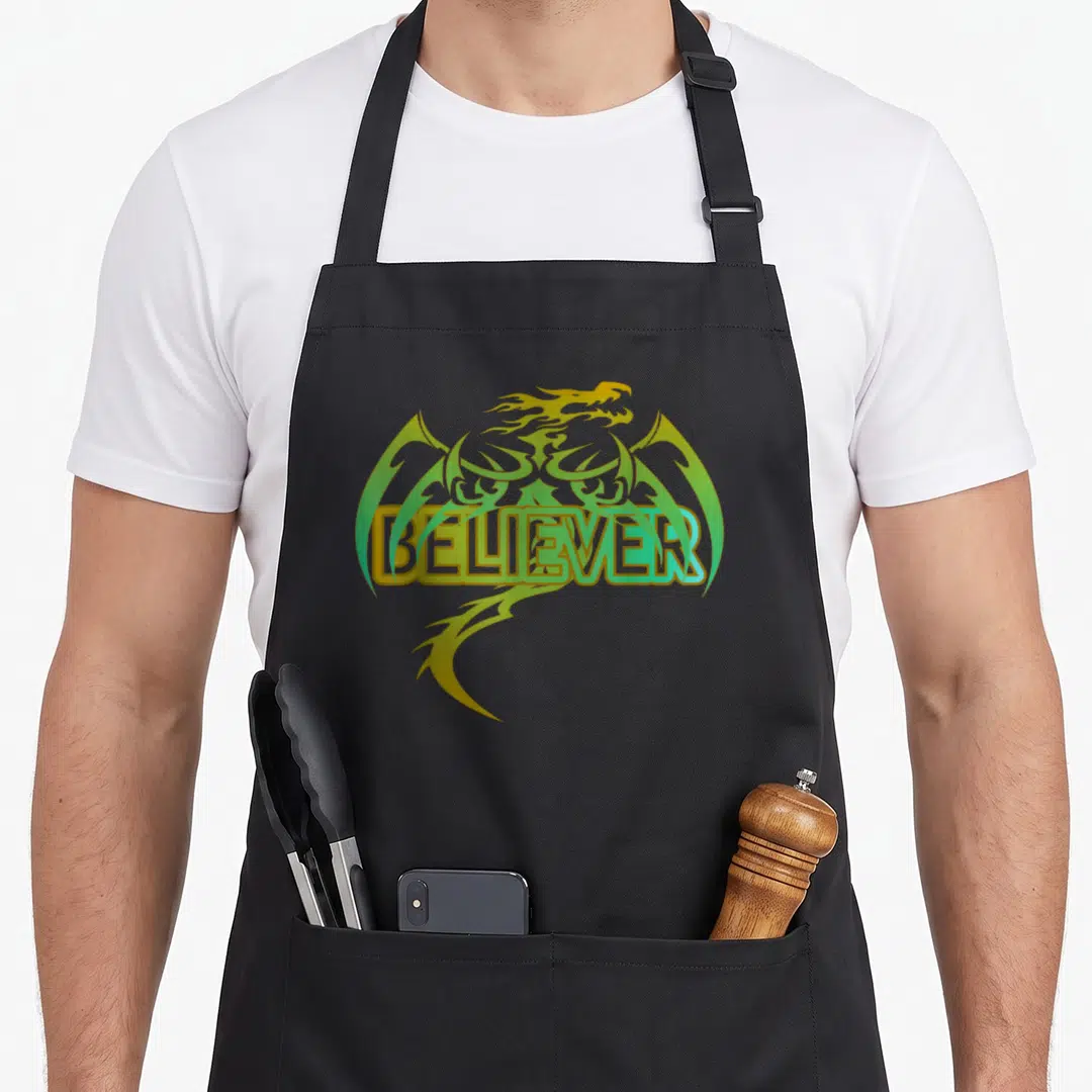 Original dragon apron - BELIEVER text over yellow and green cartoon dragon head and tail — shown on a black cotton canvas apron against a white background