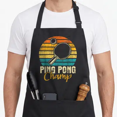 Champ table apron - Retro ping pong paddle over striped orange yellow teal sunset circle with PING PONG Champ text — shown on a black cotton canvas apron against a white background