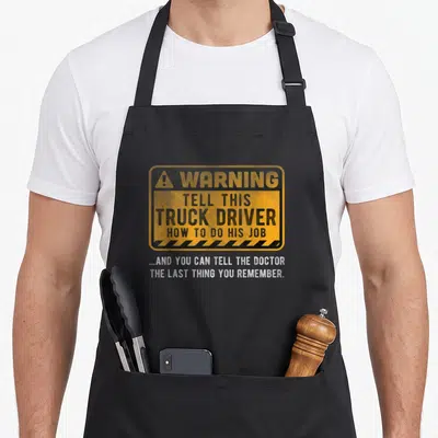 Warning truck apron - Yellow warning sign with black text and white slogan below reading about truck drivers — shown on a black cotton canvas apron against a white background
