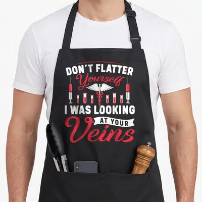 Flatter nurse apron - Don't Flatter Yourself I Was Looking At Your Veins text with caduceus and syringes — shown on a black cotton canvas apron against a white background