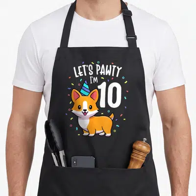 10th corgi apron - Cartoon corgi with party hat and Let's Pawty I'm 10 text with confetti — shown on a black cotton canvas apron against a white background
